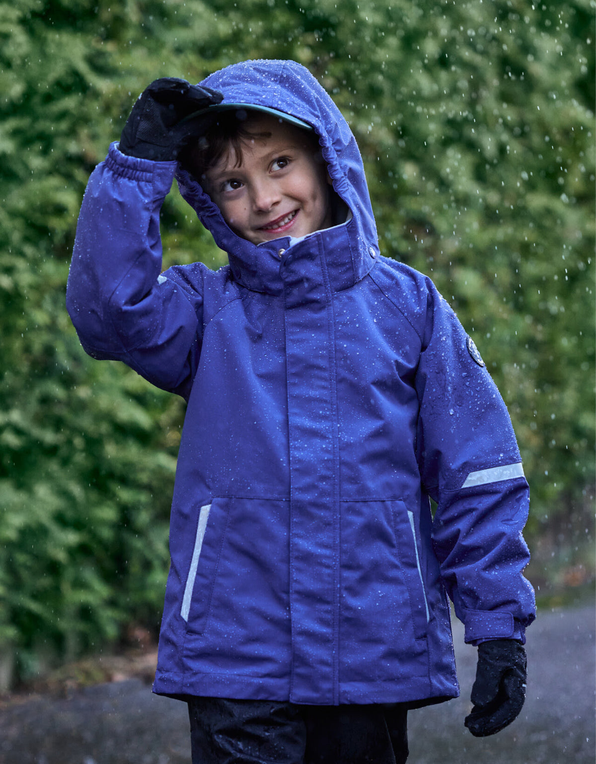A child wearing a bright blue waterproof hooded jacket and black gloves, standing in the rain outdoors with a blurred green hedge background.