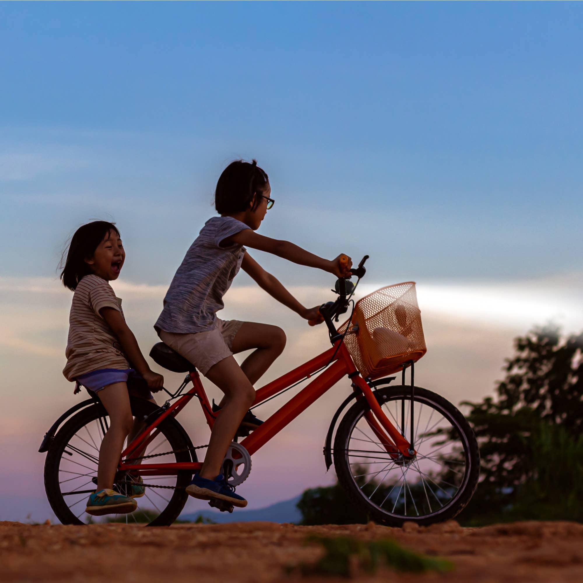 Two children riding a red bicycle together at sunset.