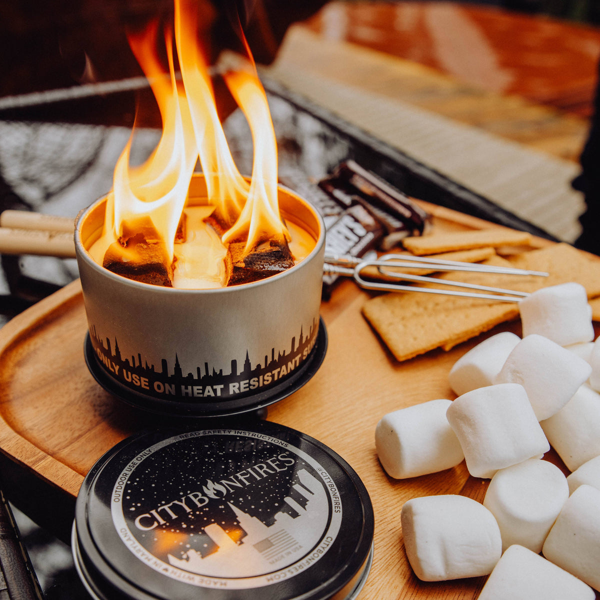 A small metal camping stove with visible flames burning inside, placed on a wooden tray with marshmallows, graham crackers, a fork, and a branded lid in front. The stove label reads 'CITY ON FIRES'.