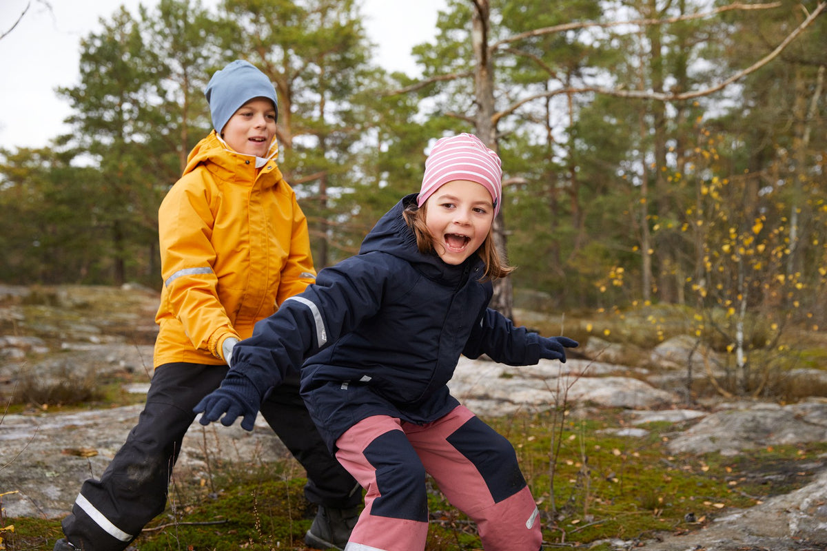 Two children outdoors in a forest setting, wearing winter clothing. The child in front wears a navy jacket, pink pants, and a pink striped beanie, while the child behind wears a yellow jacket, black pants, and a blue beanie.