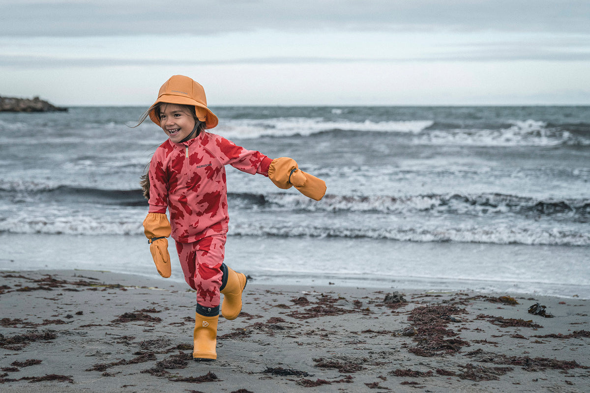 Child wearing a red camouflage rain suit, orange hat, yellow gloves, and yellow rain boots runs happily on a wet sandy beach with the ocean waves in the background.