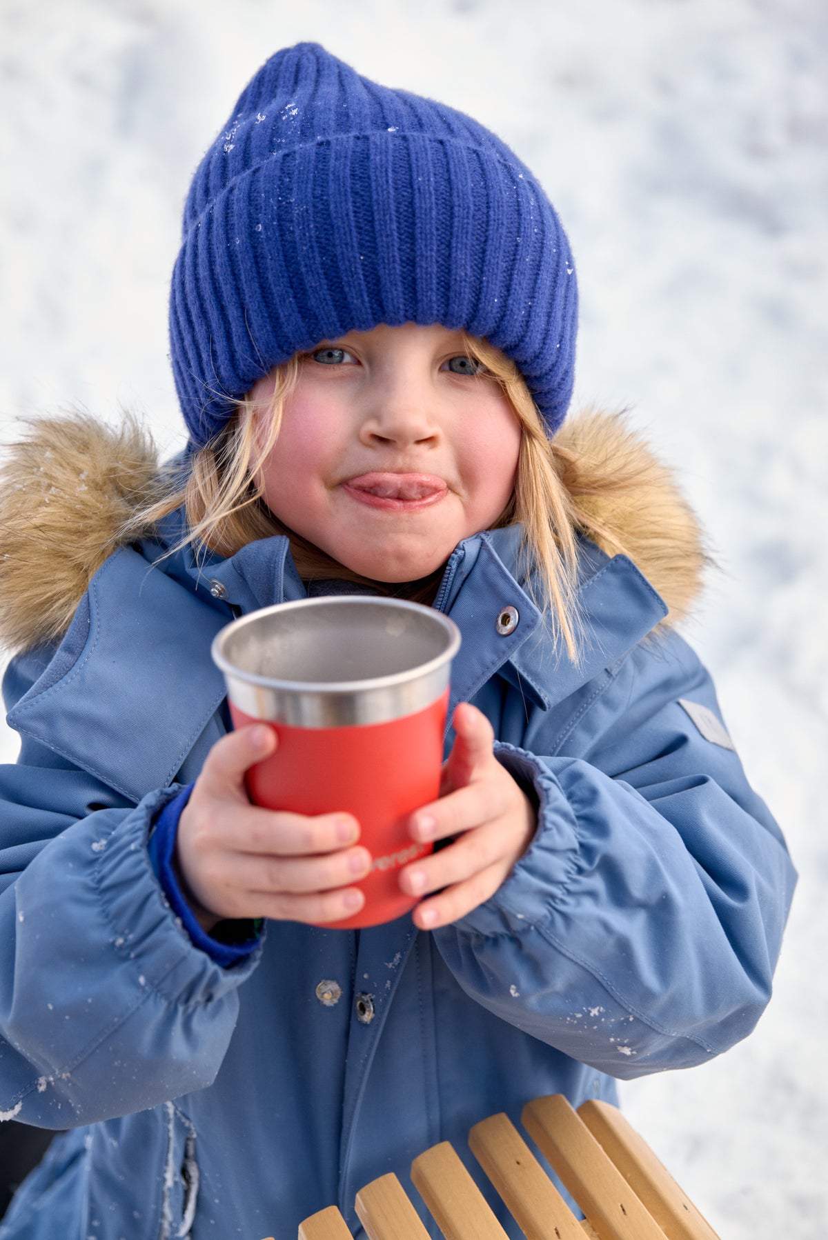 Child in winter clothing holding a red cup against a snowy background