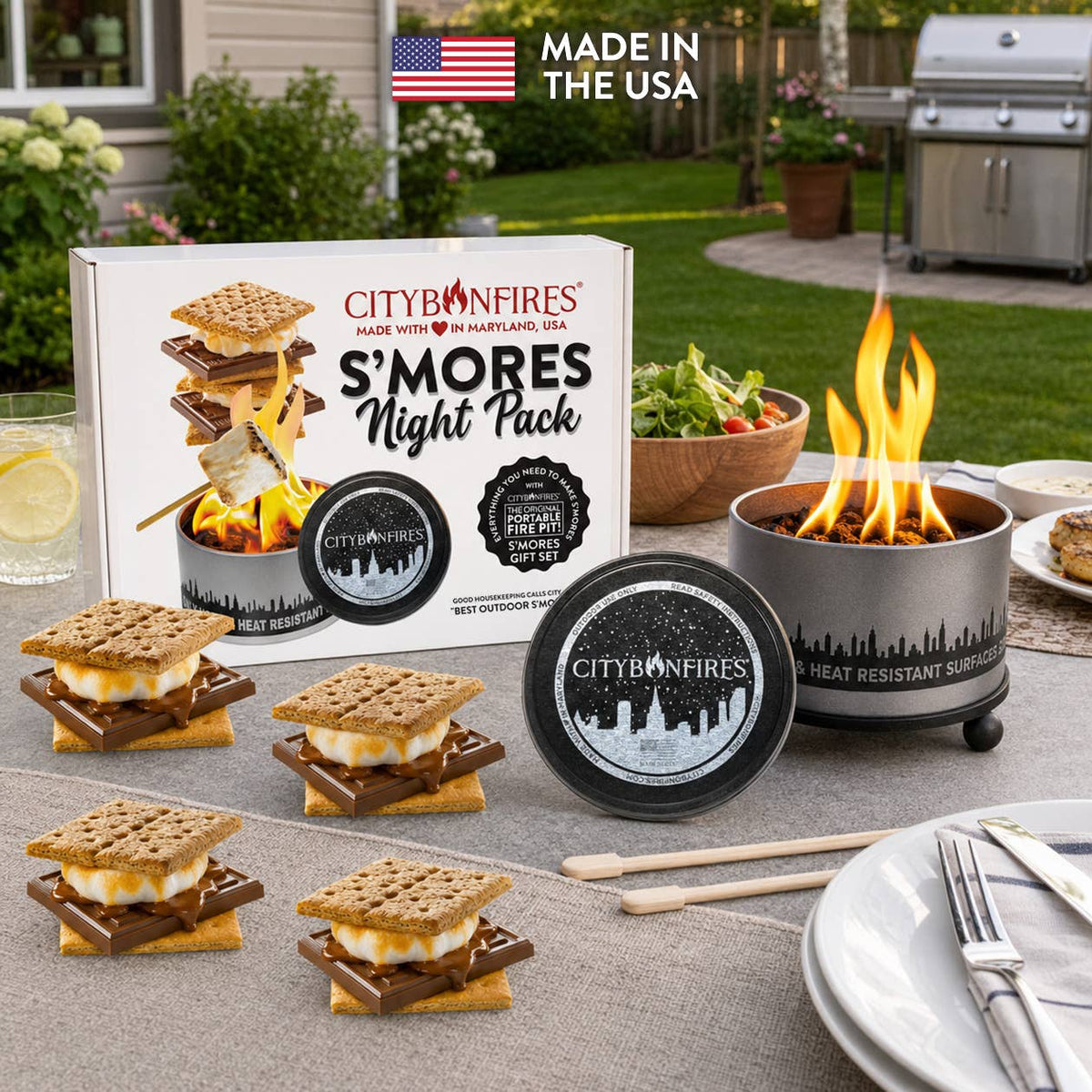 City of Fires S'mores Night Pack set displayed outdoors on a patio table, including the product box, a heat resistant s'mores burner with an active flame, a lid, several s'mores, and marshmallow sticks. A 'Made in the USA' banner is visible in the top left corner.