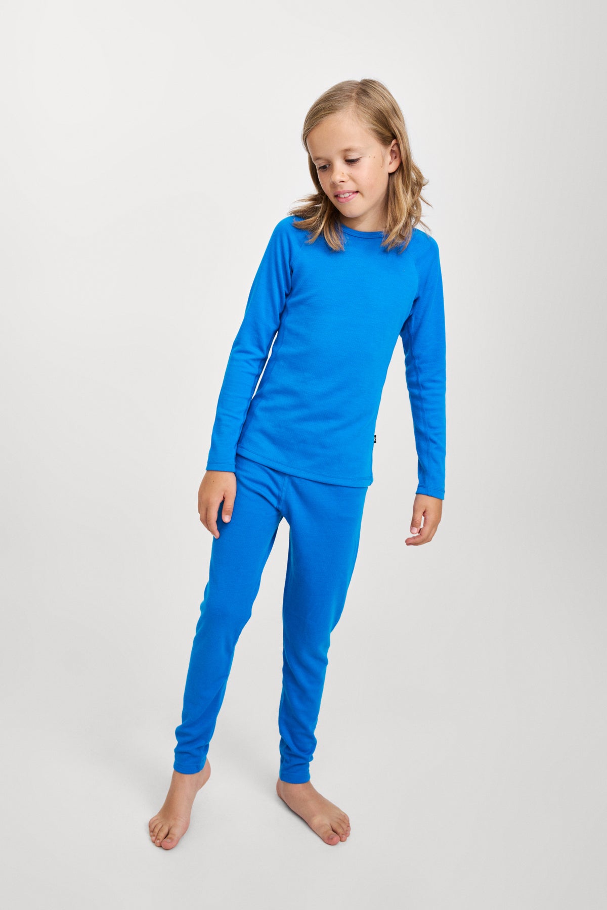 Child with blonde hair stands barefoot against a light gray studio background, wearing a bright blue long-sleeve top and matching blue pants.
