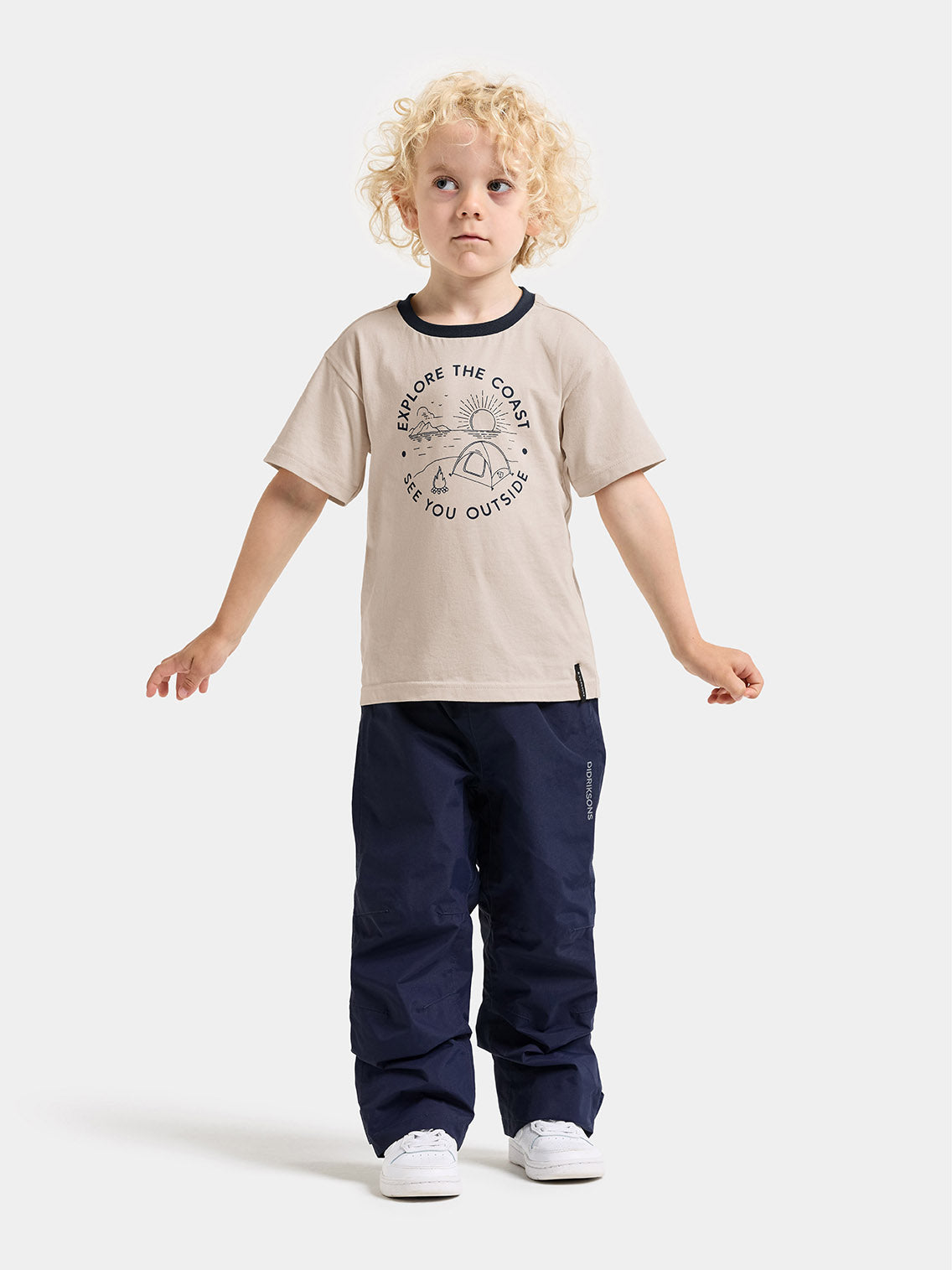 Child with curly blonde hair stands against a light gray studio background wearing a beige short-sleeve t-shirt with a dark navy graphic print, dark navy pants, and white sneakers.