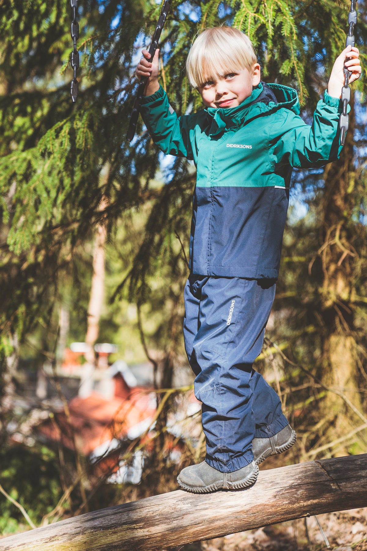Child wearing a green and navy Columbia outdoor rain suit stands on a wooden log in a forest, holding onto hanging chains.