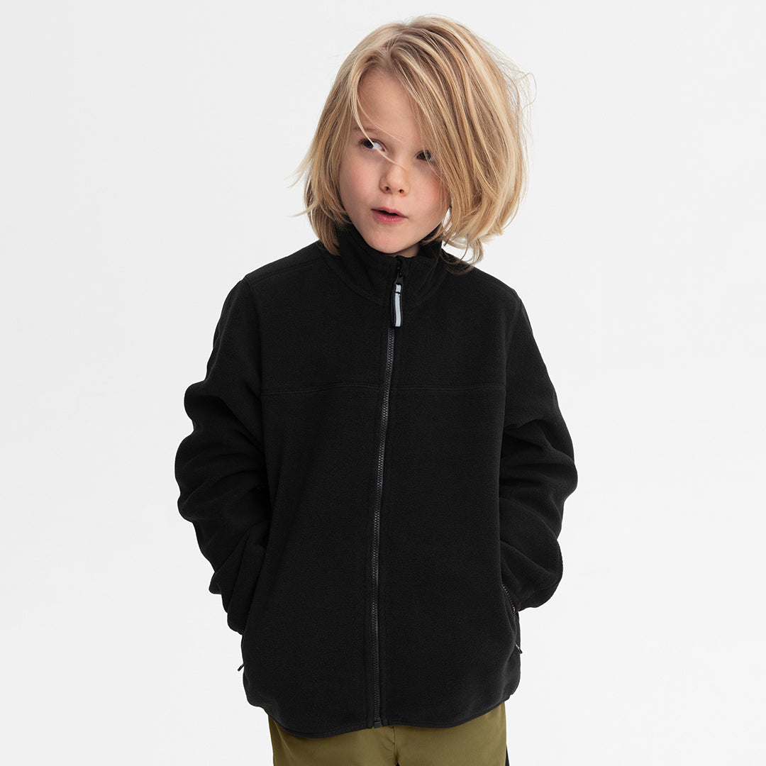 Child with blonde hair wearing a black zip-up fleece jacket and olive green pants, photographed against a clean white studio background.
