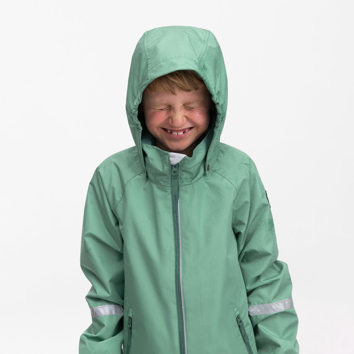 A smiling child wearing a sage green hooded rain jacket with reflective silver stripes on the sleeves, photographed against a clean white background.