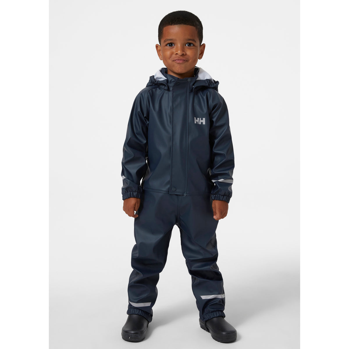 Child wearing a navy blue waterproof hooded rain suit with reflective silver stripes and the Helly Hansen logo on the chest, standing against a light gray studio background.