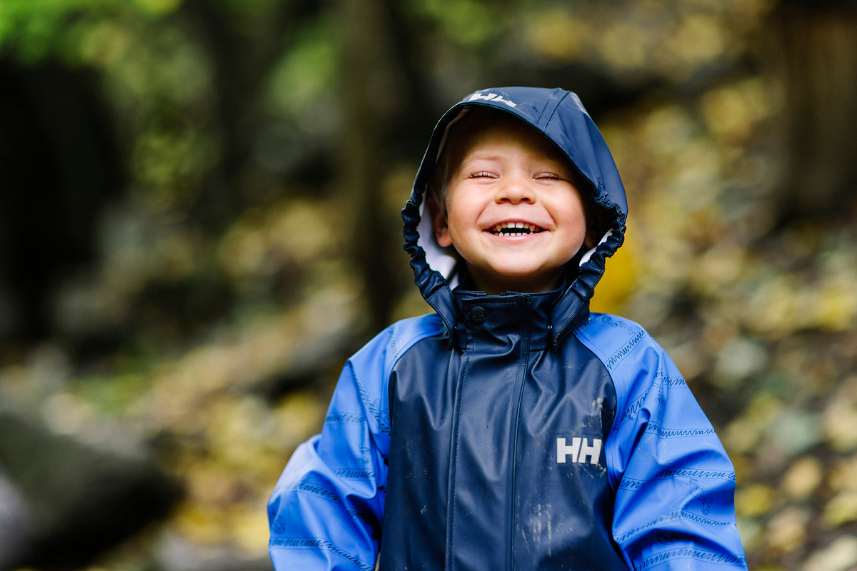 A young boy wearing a blue and navy Helly Hansen rain jacket with the hood up, smiling outdoors in a forest setting.
