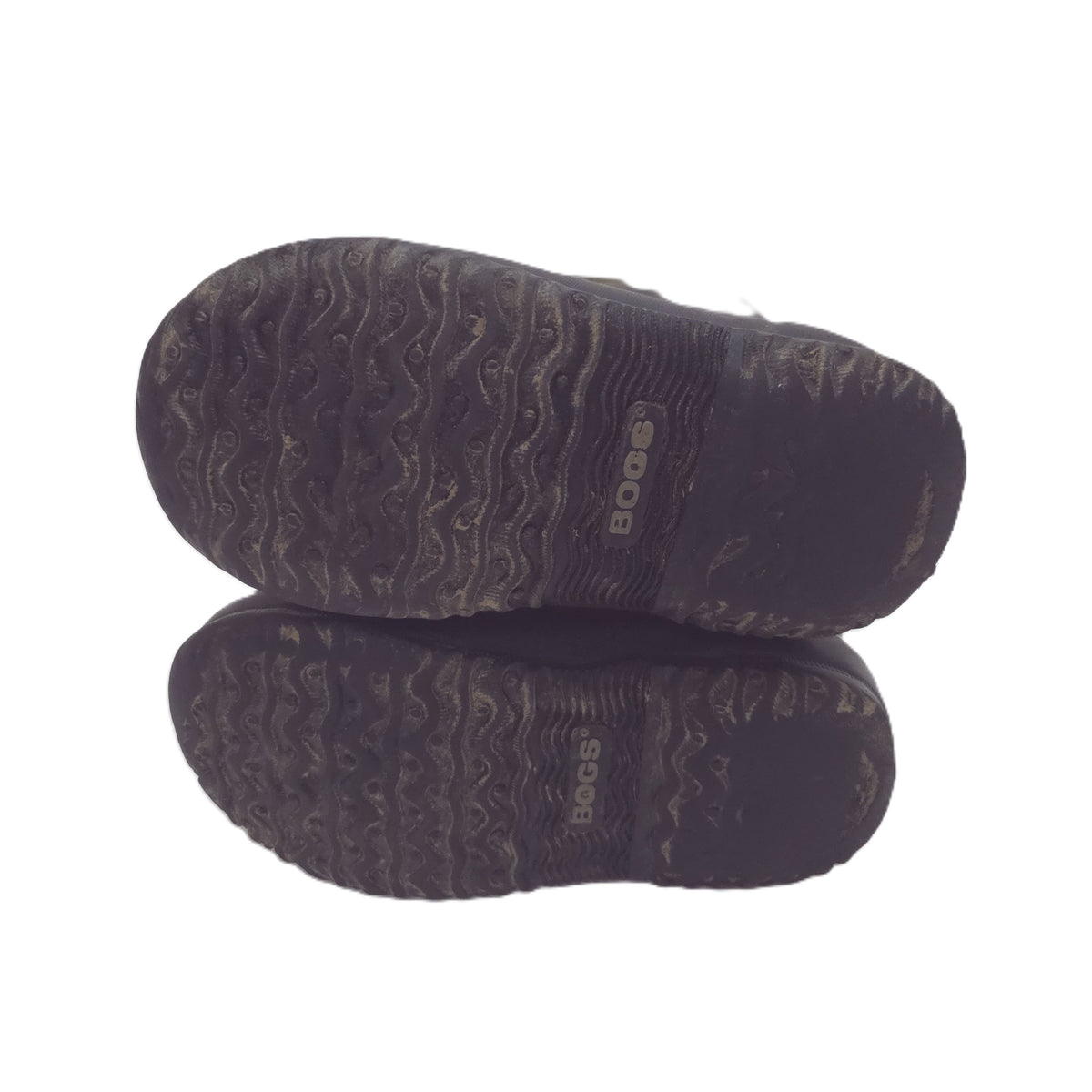 A pair of black outdoor boots shown from the bottom, displaying their textured wavy tread soles with the brand name 'BOGS' printed in white on the heel area, set against a plain white background.