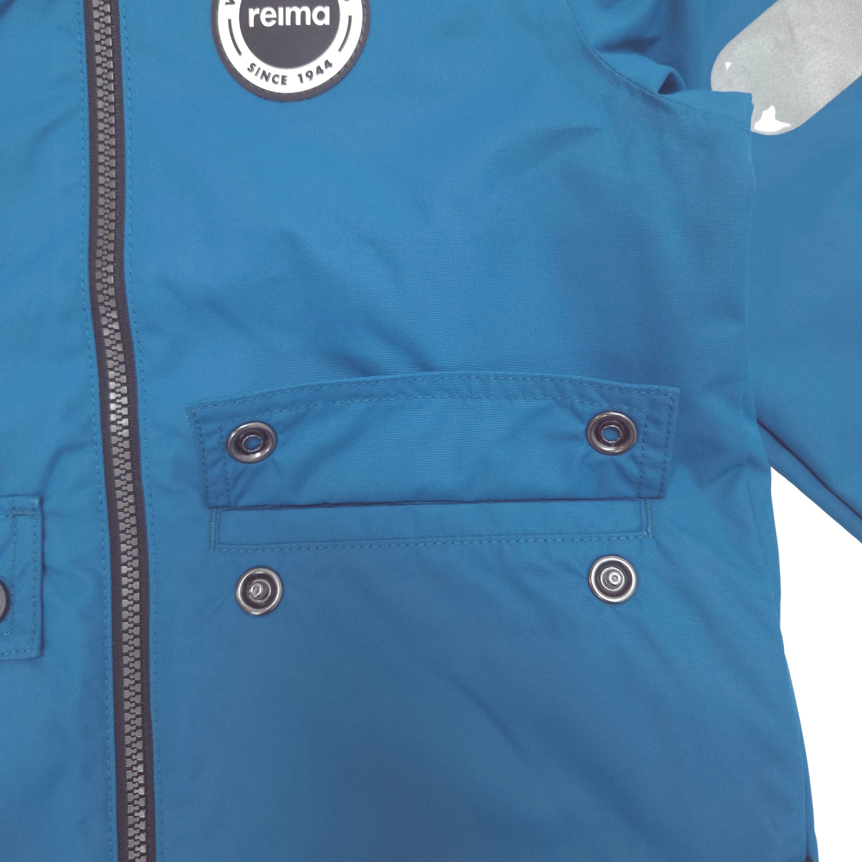 Close-up of a blue Reima jacket showing a front zipper, a hand pocket with snap buttons, and a circular Reima logo patch on the upper chest.