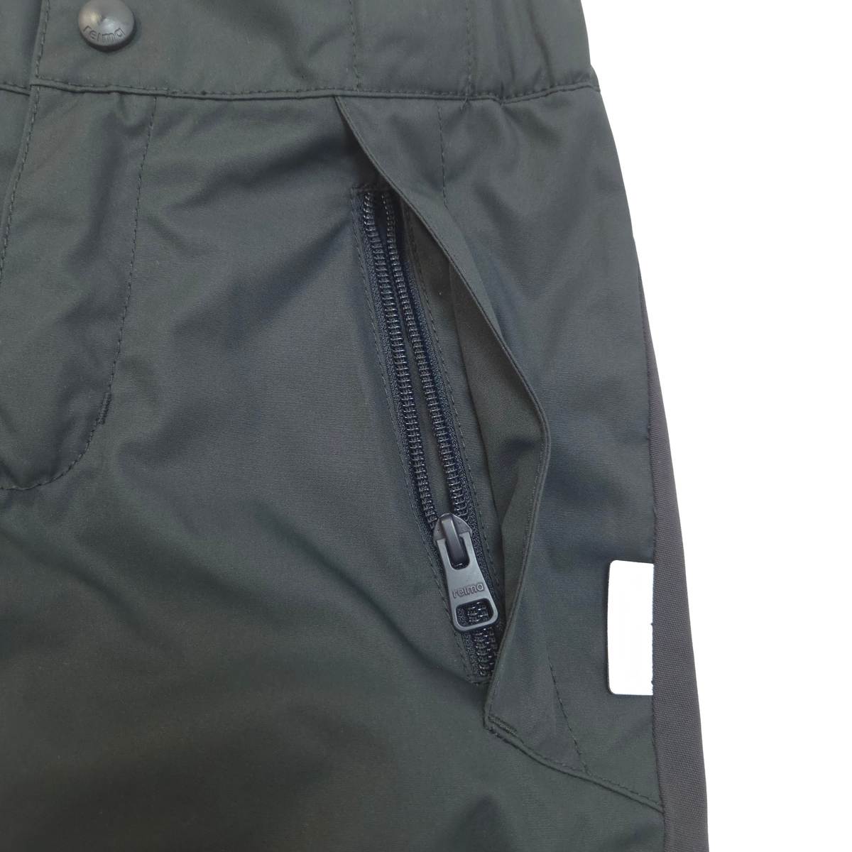 Close-up of black outdoor pants showing a zippered pocket, button waist, and a small white rectangular tag.