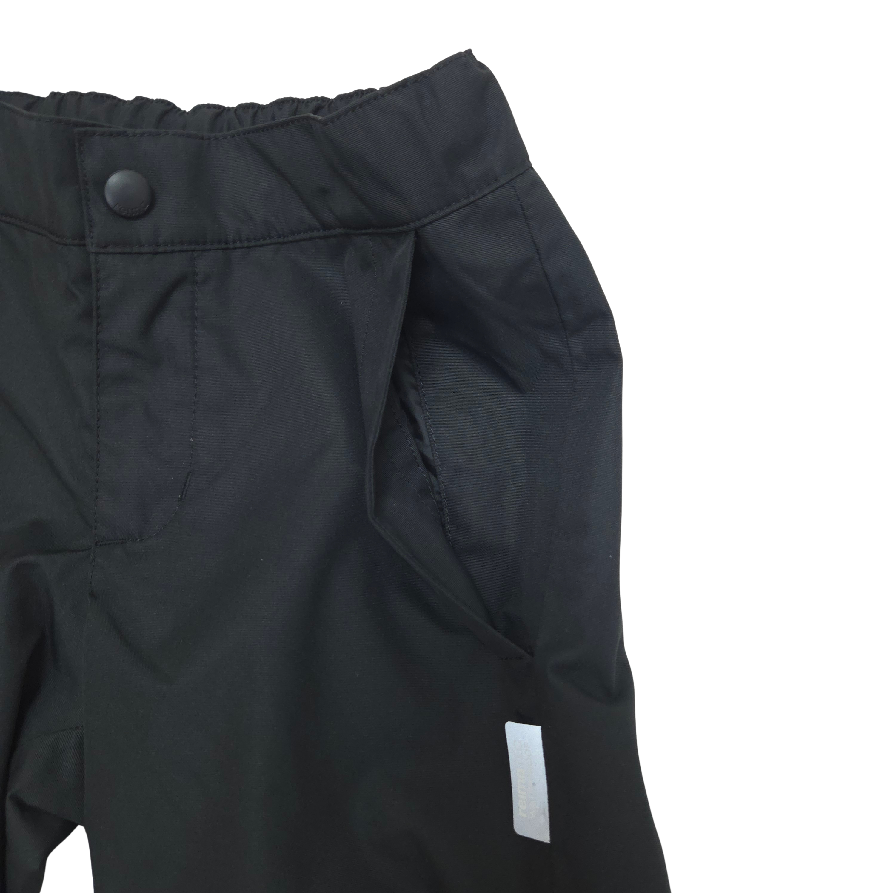 Close-up of black outdoor pants showing the waistband with a snap button, side pocket, and a small white rectangular reflective tag on a plain white background.
