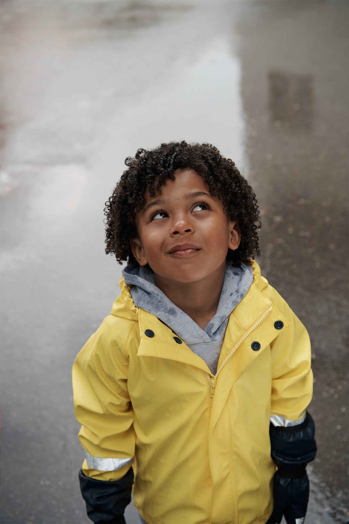 Child looks upward with a thoughtful expression, wearing a bright yellow rain jacket over a gray hoodie and black gloves, standing outdoors on a wet gray pavement.