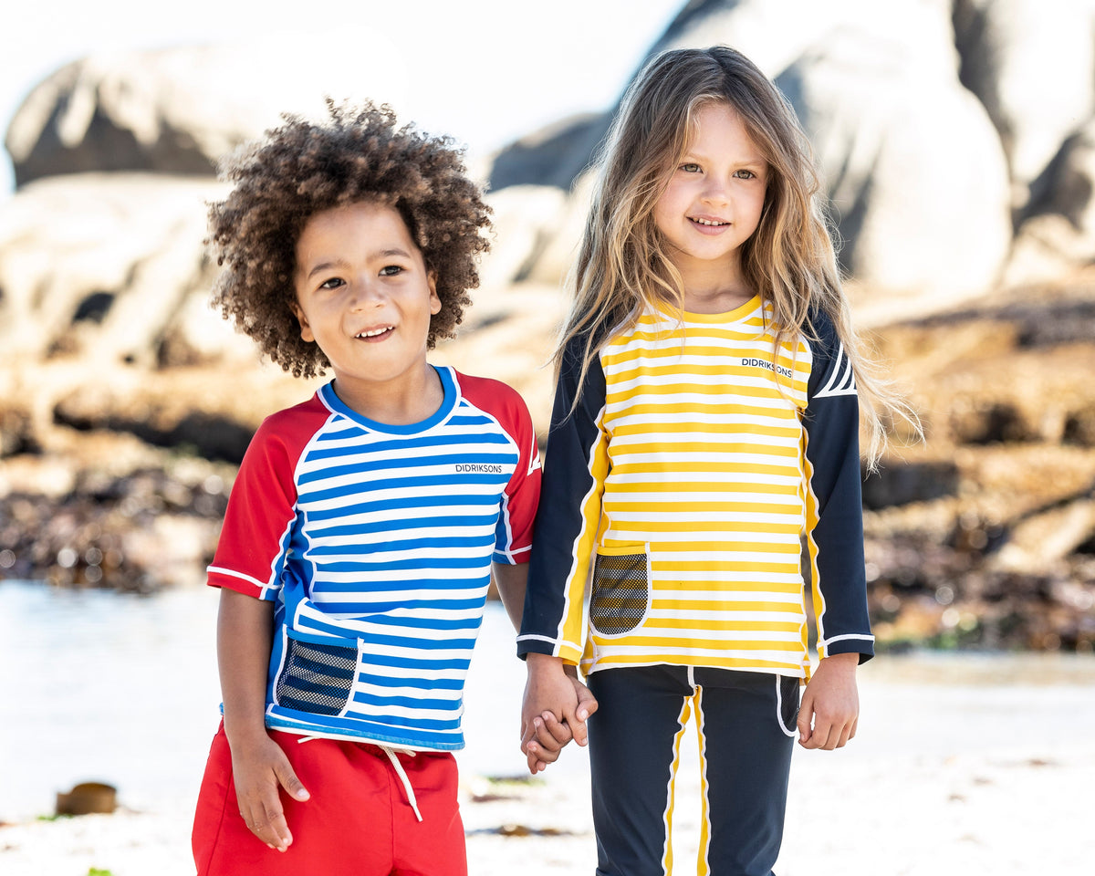 Two children standing on a sandy beach holding hands, wearing matching striped rash guard swim shirts. One child wears a blue and white striped shirt with red sleeves and red swim trunks, while the other wears a yellow and white striped shirt with navy sleeves and navy swim pants with yellow stripes.