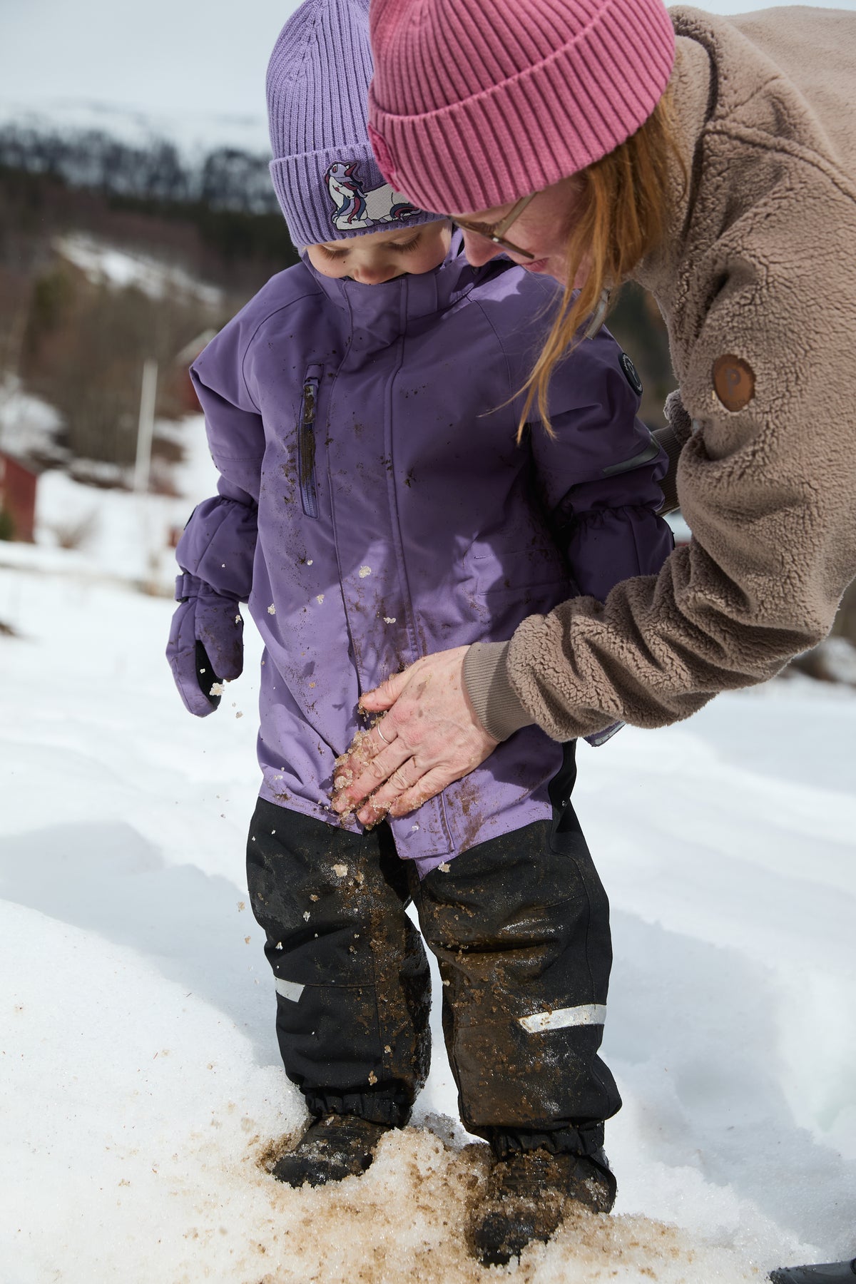 Child with muddy winter clothing and adult in pink hat standing in the snow.