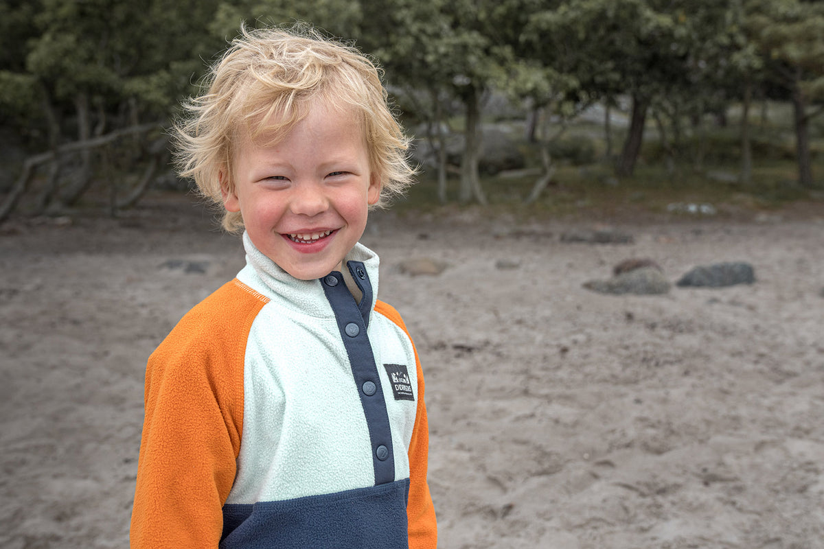 Child smiling outdoors on a sandy beach, wearing a color-blocked fleece pullover with orange, off-white, and navy panels.