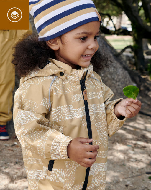Child wearing a beige jacket and striped hat holding a leaf outdoors