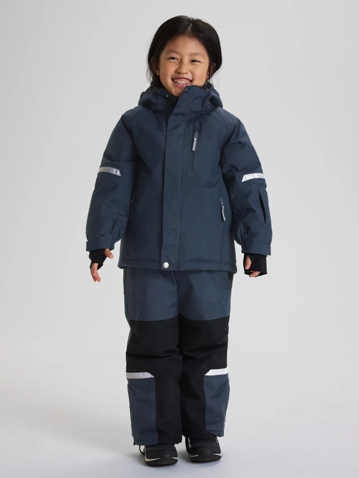 A young child wearing a dark navy blue Polarn O. Pyret winter jacket and matching pants with reflective light gray stripes, standing against a light gray studio background.