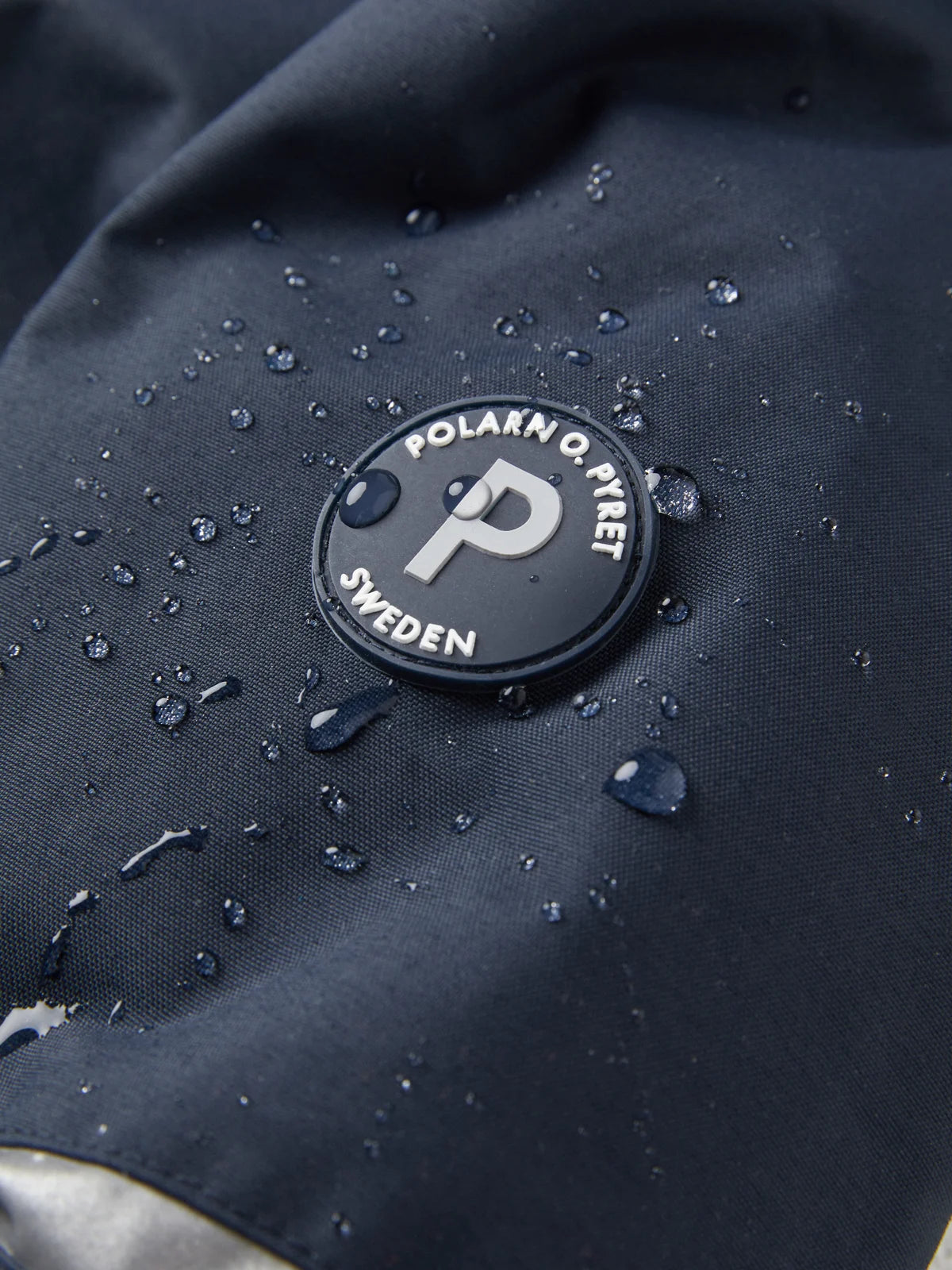 Close-up of a black circular Polarn O. Pivet Sweden logo patch on a dark navy waterproof fabric, with water droplets beaded on the surface showing the inherent waterproof nature of this jacket.
