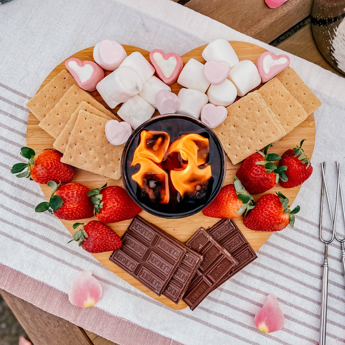 Heart-shaped charcuterie board with strawberries, marshmallows, graham crackers, and chocolate on a striped tablecloth.