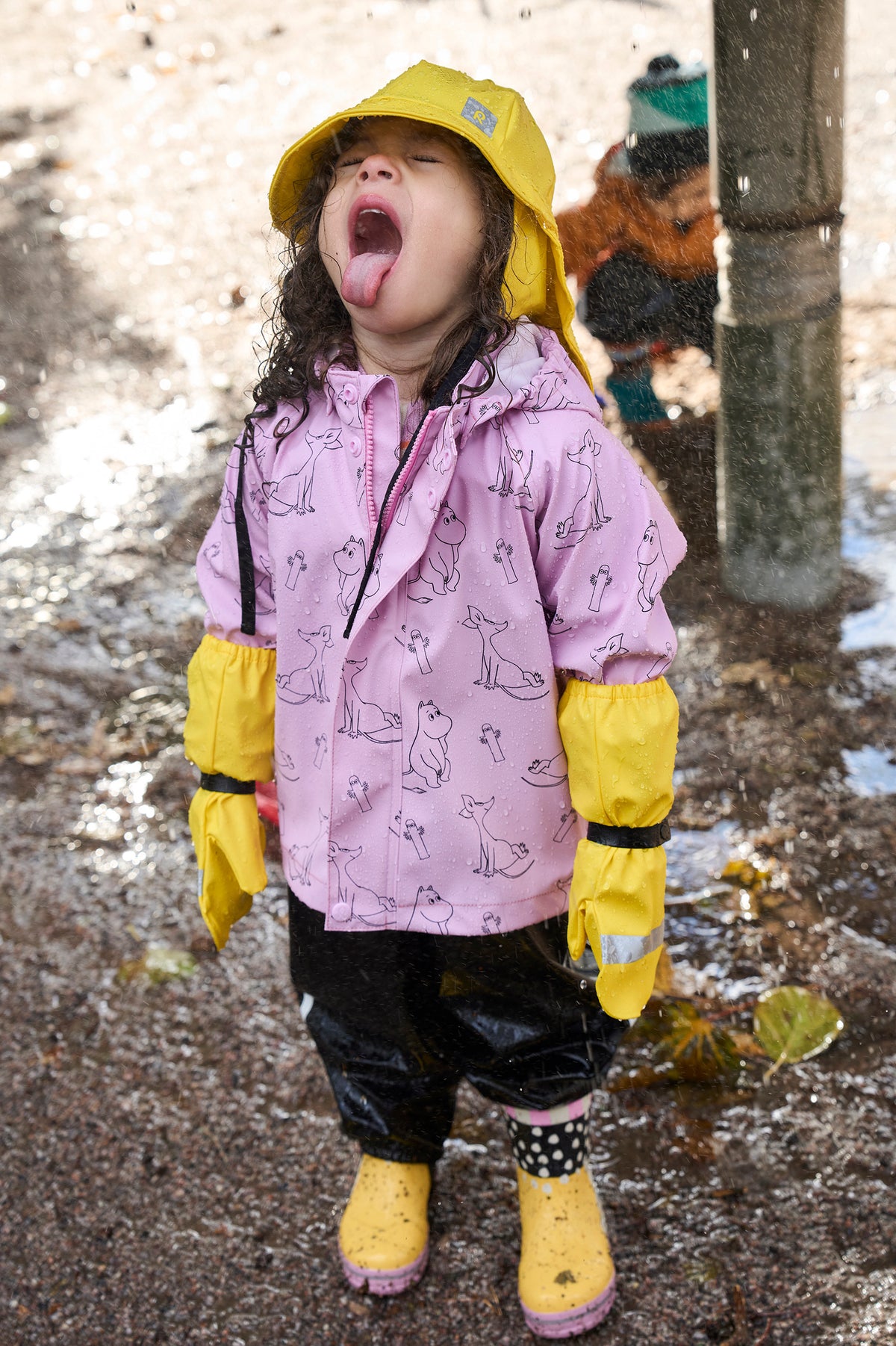 A young child wearing a pink raincoat with a cat pattern, yellow rain hat, yellow rain boots, and yellow rain sleeves, sticking their tongue out in a puddle outdoors.