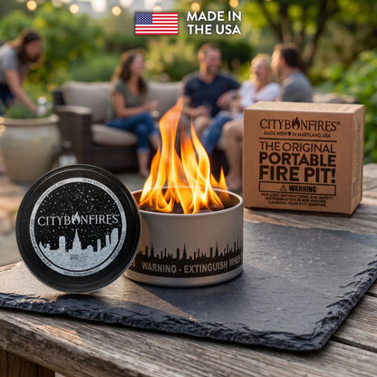 A portable fire pit set on a wooden table outdoors, with a small metal fire pit showing flames, its lid placed beside it, and the branded box behind it. A blurred patio seating area and people are visible in the background, along with a 'Made in the USA' badge at the top.