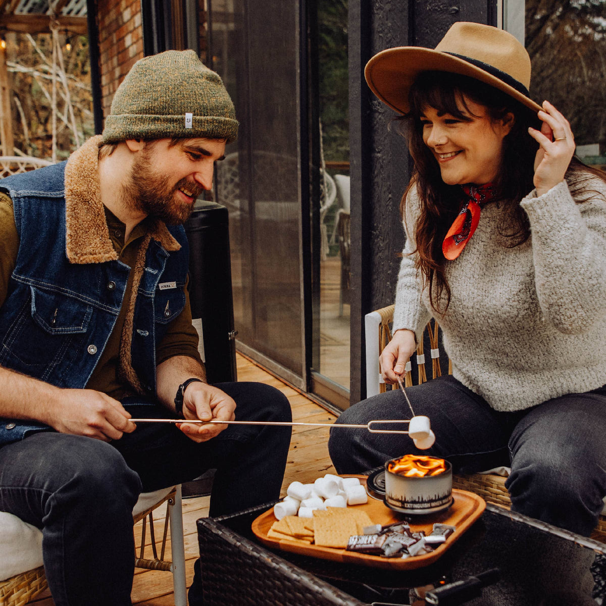 A man and a woman sit on a wooden deck chair by a window, roasting marshmallows over a small campfire. The man wears a green beanie, denim vest, and brown shirt, while the woman wears a wide-brim tan hat, gray knit sweater, and red neck scarf.