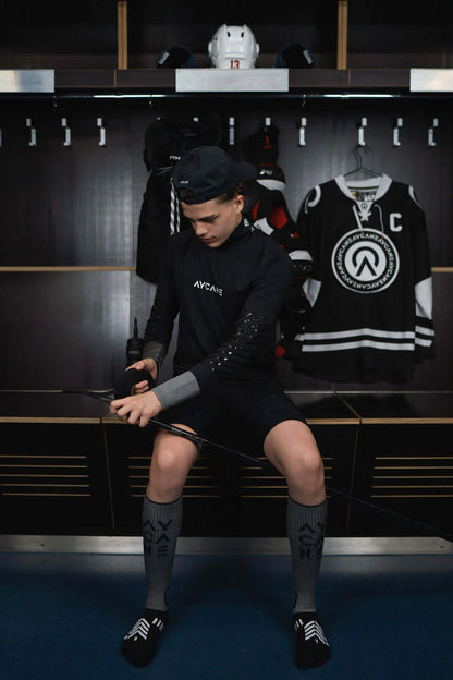 A hockey player sitting in a locker room wearing a black long-sleeve performance jersey with 'AYCANE' on the chest, black shorts, and gray knee-high socks.
