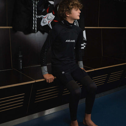 A young boy wearing a black long-sleeve athletic shirt with 'AVEAME' printed on the chest and matching black leggings, sitting on a bench in a locker room.