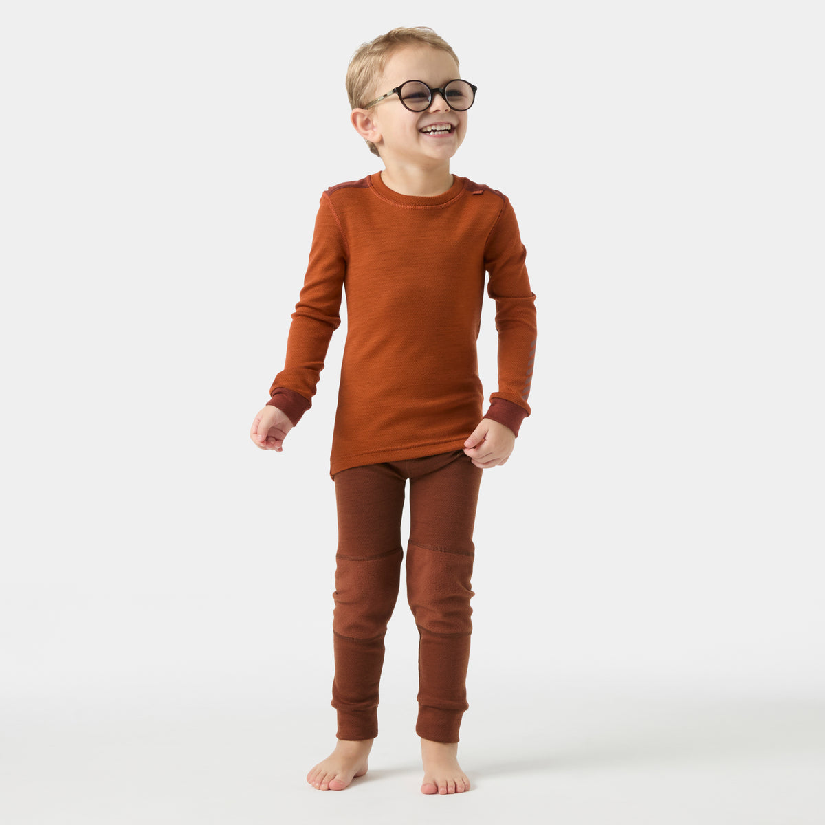 Child wearing a matching rust-orange long-sleeve top and brown pants, standing barefoot against a plain light gray studio background.