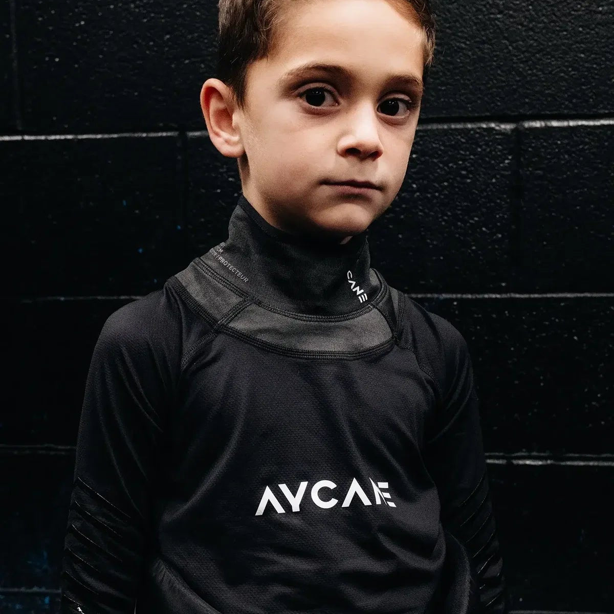 A young boy wearing a black long-sleeve athletic shirt with a high collar and white 'AYCANE' text on the chest, standing against a dark textured wall.