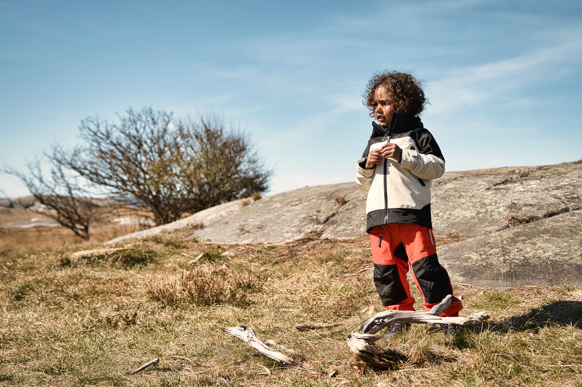 A child stands outdoors on a grassy hillside under a blue sky, wearing a black and off-white zip jacket and bright red pants.