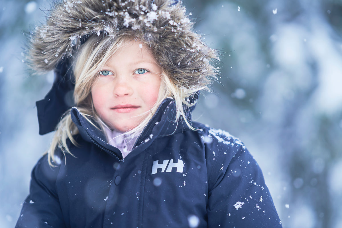 Child wearing a navy blue Helly Hansen winter jacket with a white HH logo on the chest, a brown fur-trimmed hat, and a light purple shirt, photographed outdoors in the snow.