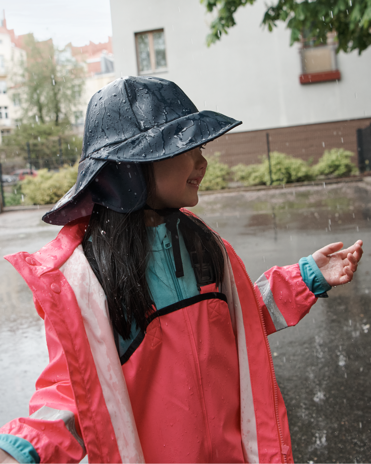 A child wearing a bright pink raincoat and a dark blue rain hat stands in the rain on a wet city street, looking to the side with one hand raised.