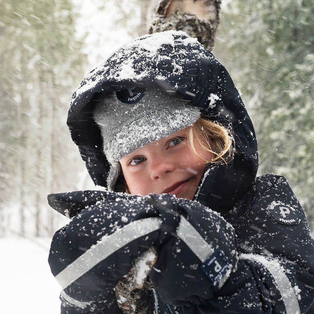 A child wearing a black winter snowsuit with reflective stripes, a gray knit hat, and a black balaclava hood covered in snow, smiling in a snowy forest.