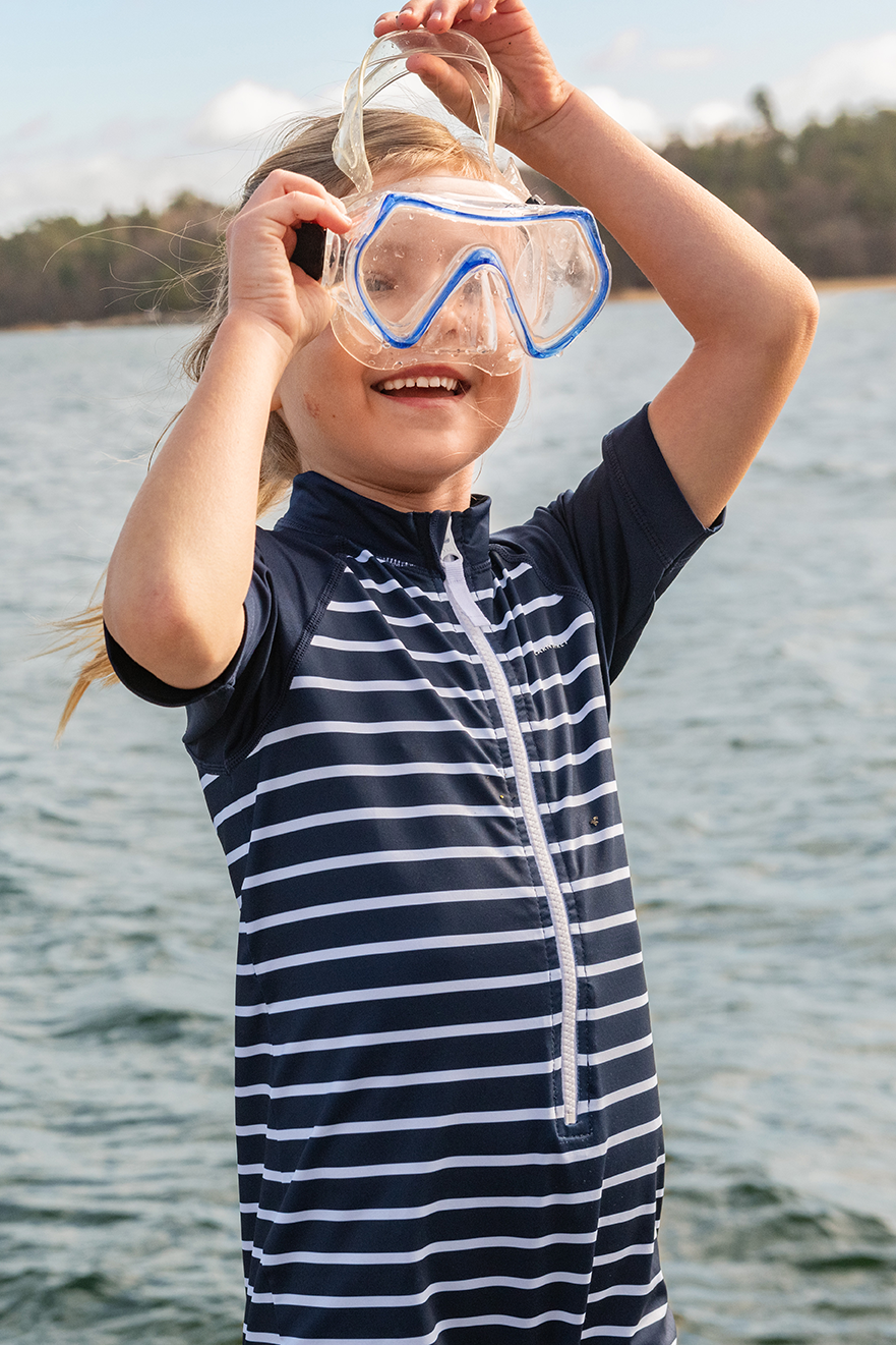 A young child wearing a navy and white striped swim shirt and a blue snorkel mask, standing outdoors by a body of water with a tree-lined shore in the background.