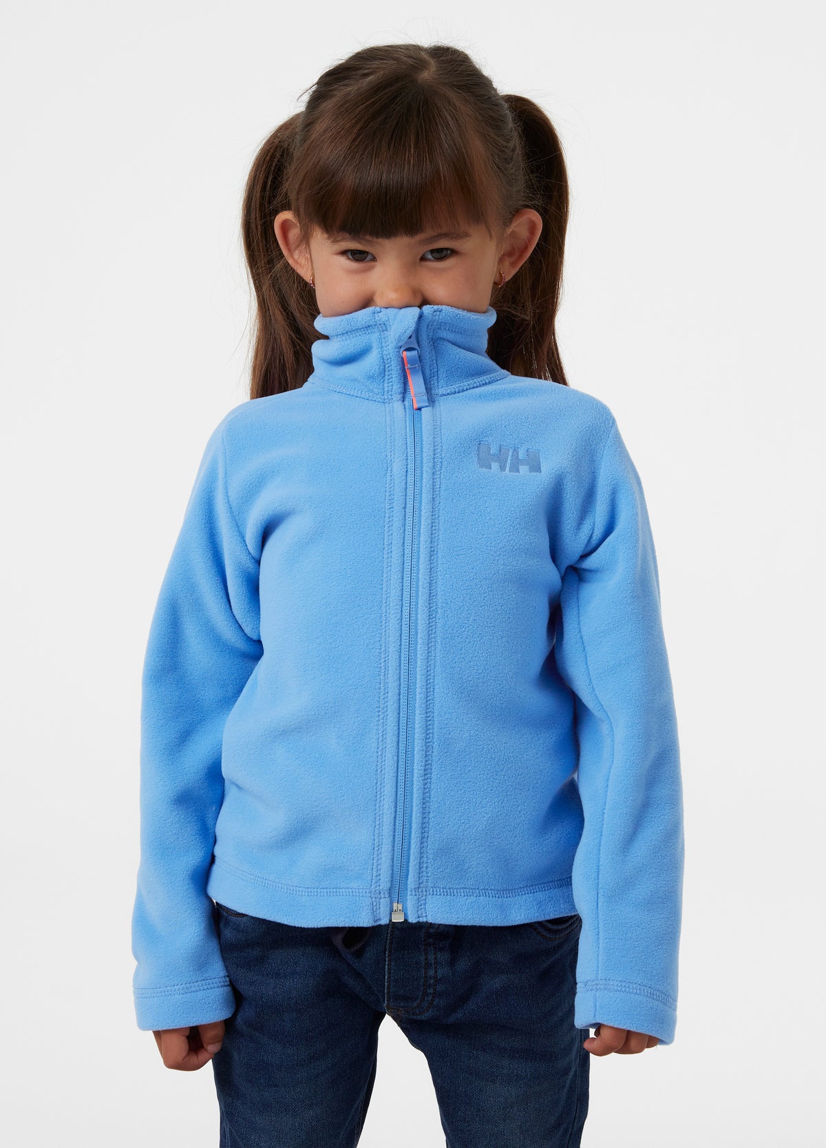 Child ith brown hair in pigtails wearing a light blue Helly Hansen zip-up fleece jacket and dark blue jeans, standing against a plain white studio background.