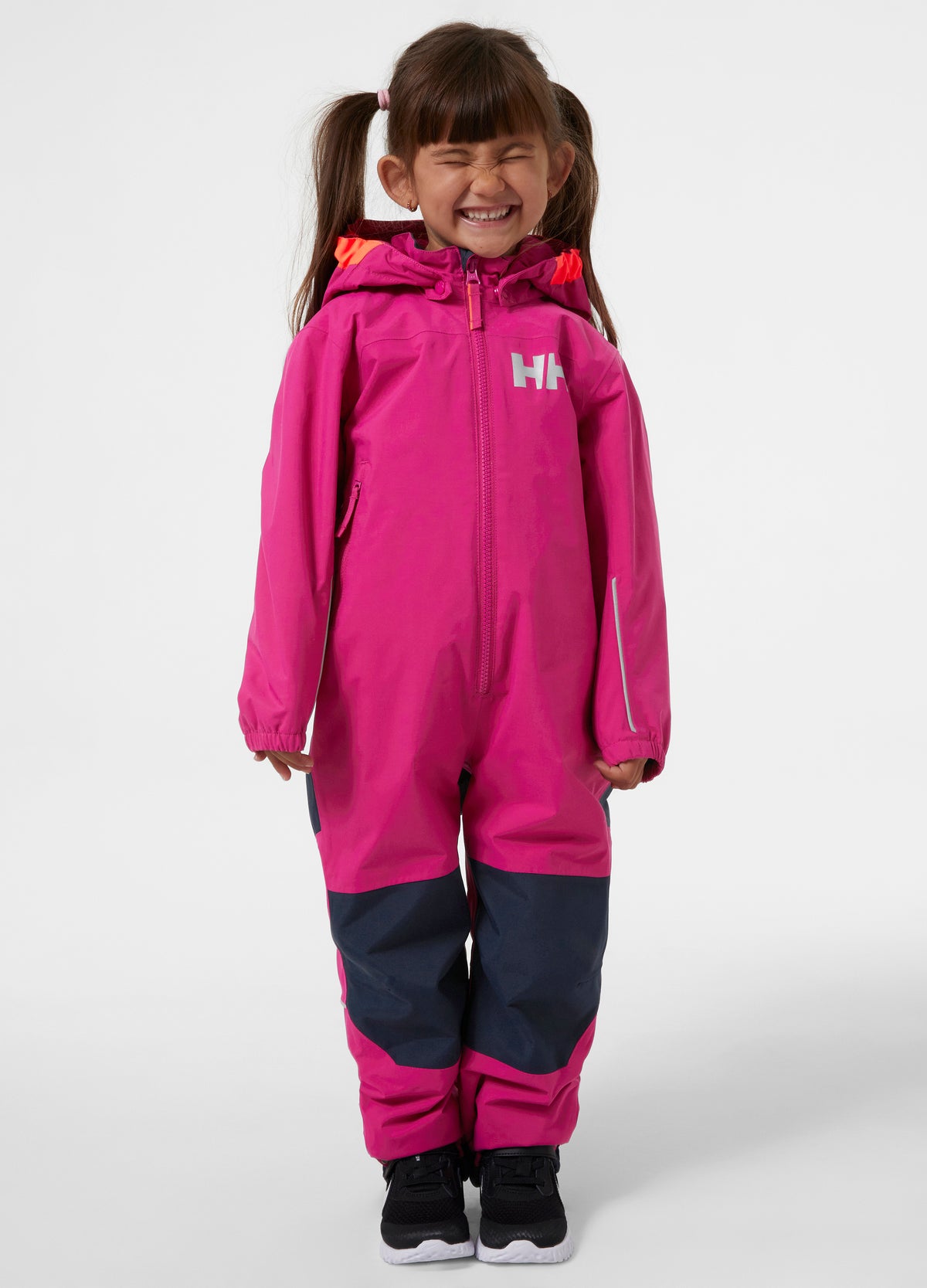 Child smiling and wearing a bright pink one-piece snowsuit with dark navy lower legs and cuffs, standing against a light gray studio background. The brand logo 'HH' is visible on the chest.
