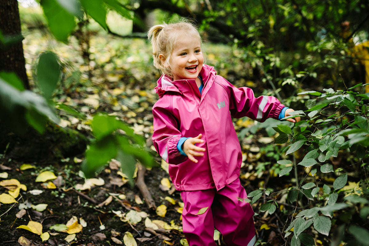 A young girl with blonde hair in a ponytail smiles and reaches out while wearing a bright pink waterproof rain suit in a lush green outdoor setting.
