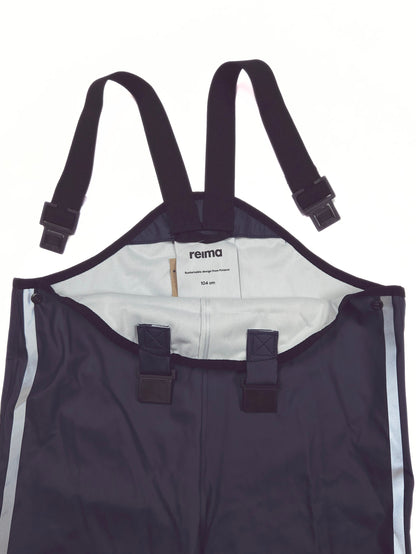 Close-up of a Reima waterproof bib , showing the dark navy exterior, adjustable black shoulder straps with plastic buckles, a white inner lining, and reflective silver side stripes.