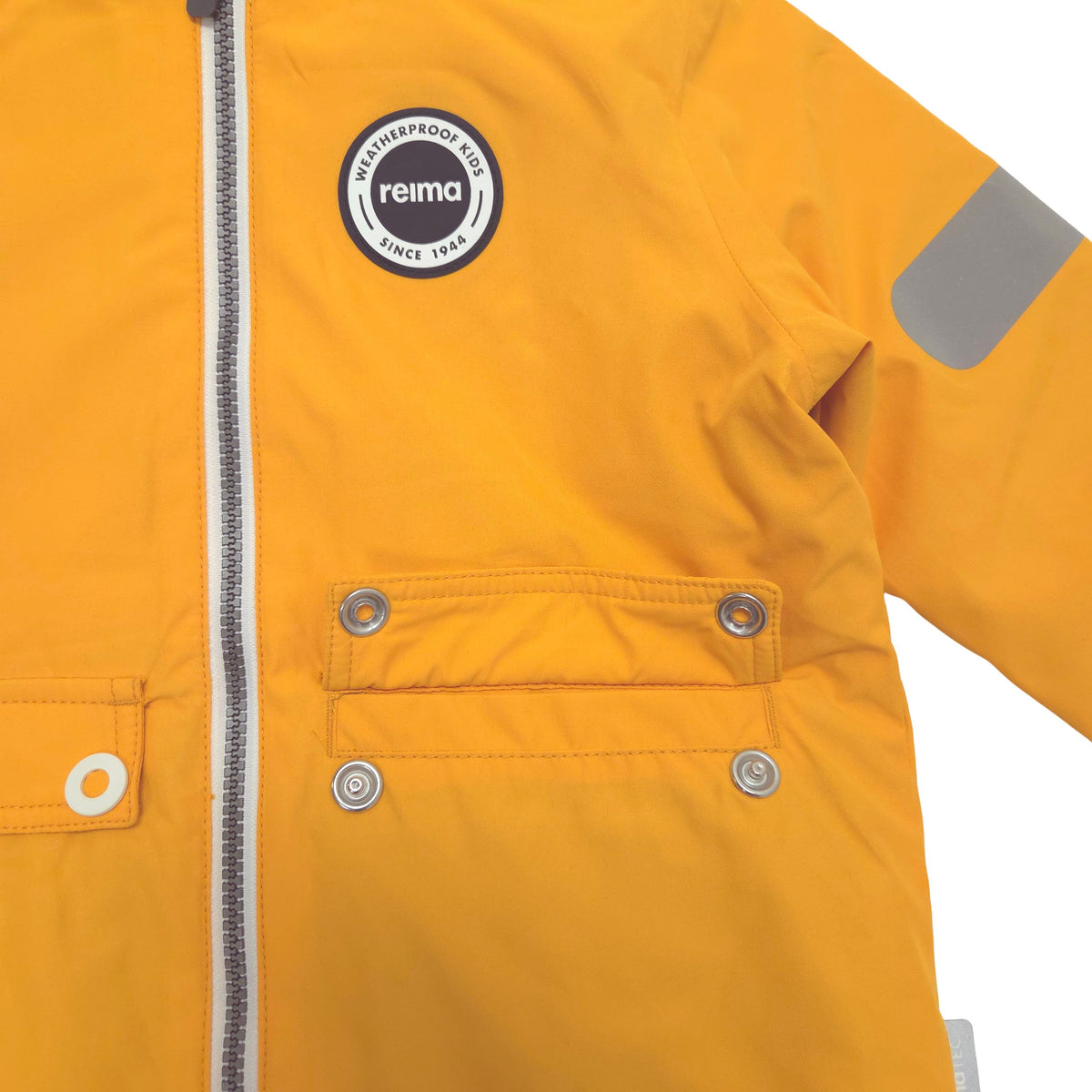 Close-up of a bright yellow waterproof jacket with a front zipper, flap pockets with metal snap buttons, a circular 'reima' logo patch on the chest, and a reflective strip on the sleeve.