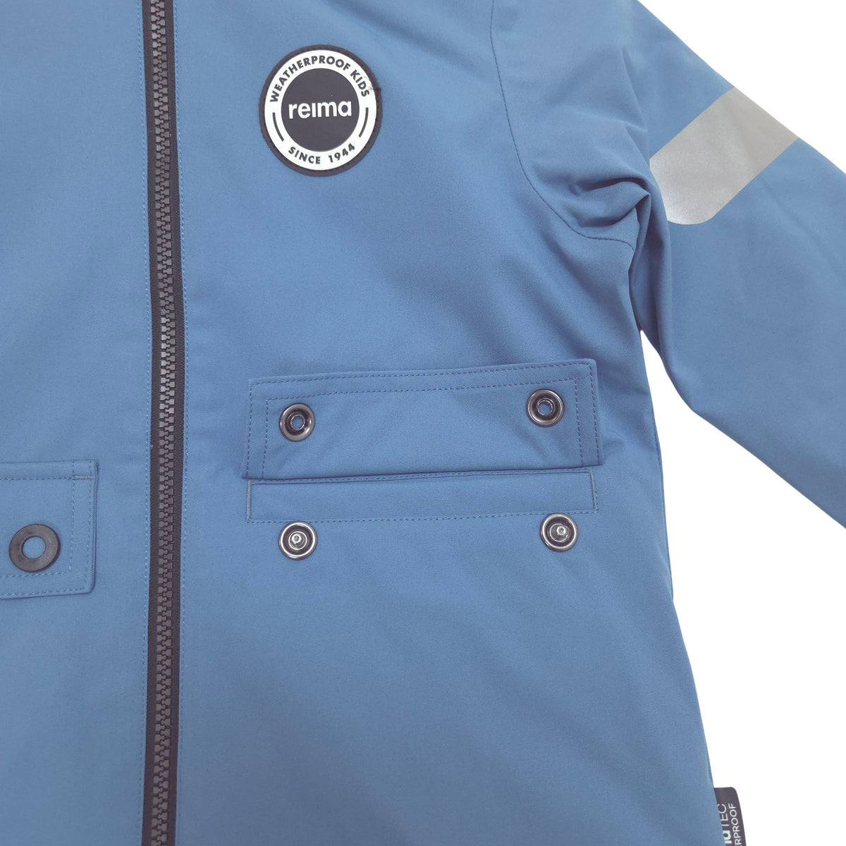 Close-up of a light blue waterproof jacket with a circular black and white Reima logo patch on the chest, a gray reflective stripe on the sleeve, and two snap flap hand pockets