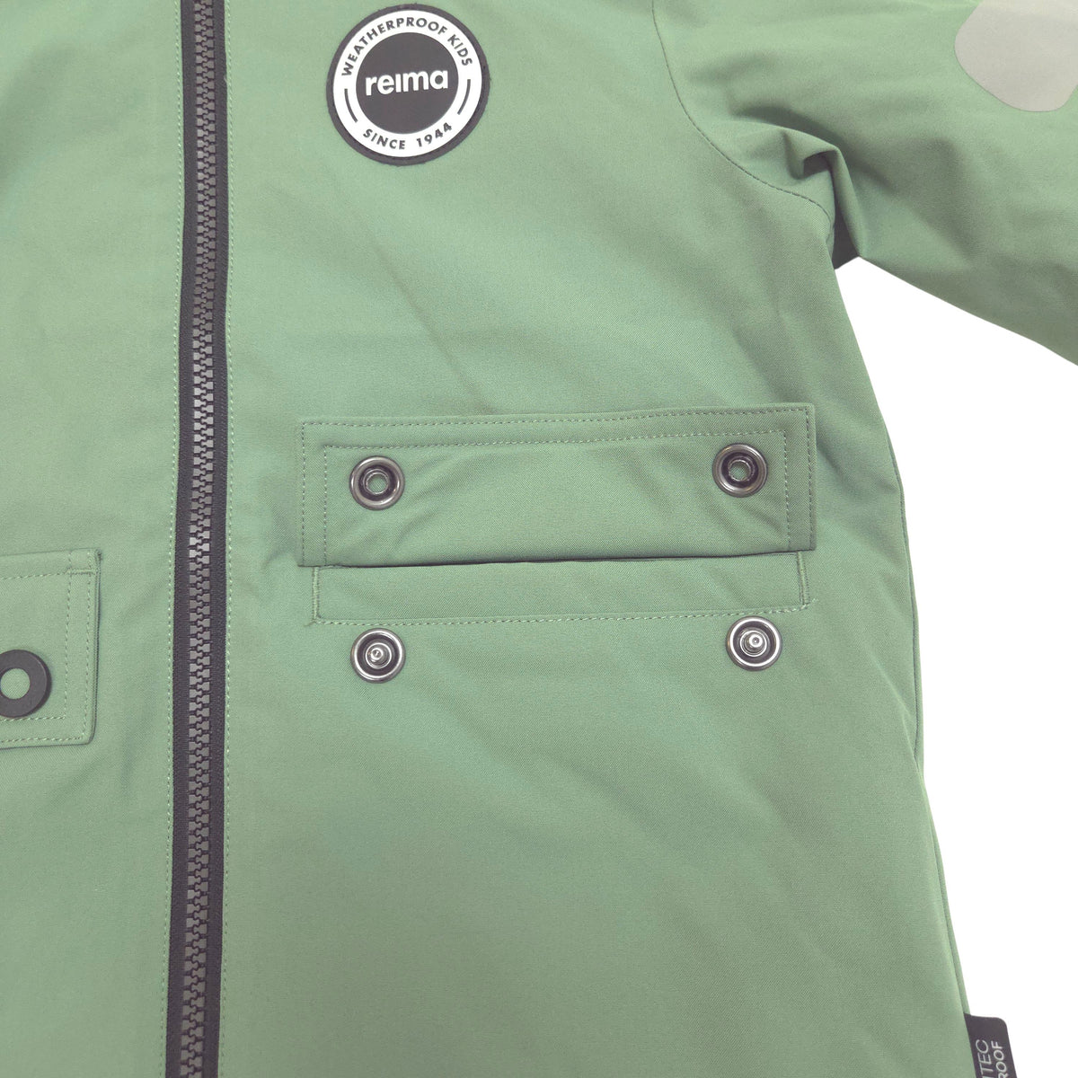 Close-up of a sage green jacket showing the chest area, including a circular 'reima' brand patch, a front zipper, and a flap pocket with snap buttons.