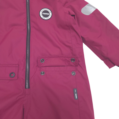 A close-up of a children's magenta pink snowsuit jacket by reima, featuring a front zipper, snapped flap pockets, and a reflective silver patch on the sleeve.