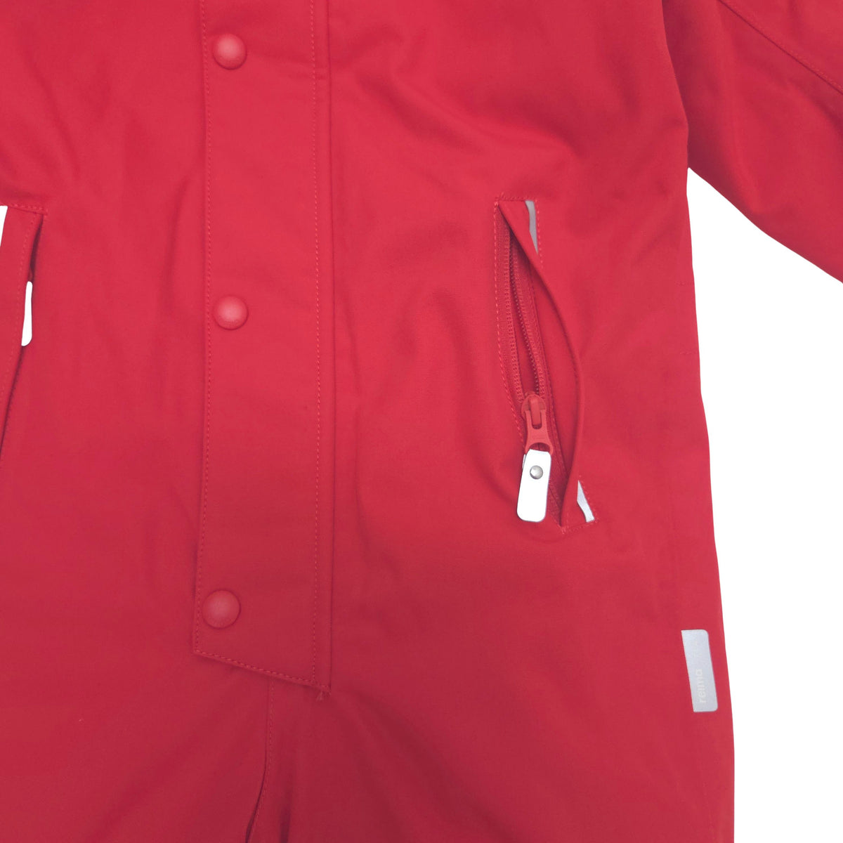 Close-up of a red snow suit showing a buttoned front flap, a zippered pocket with a reflective zipper pull, and a small reflective tab.