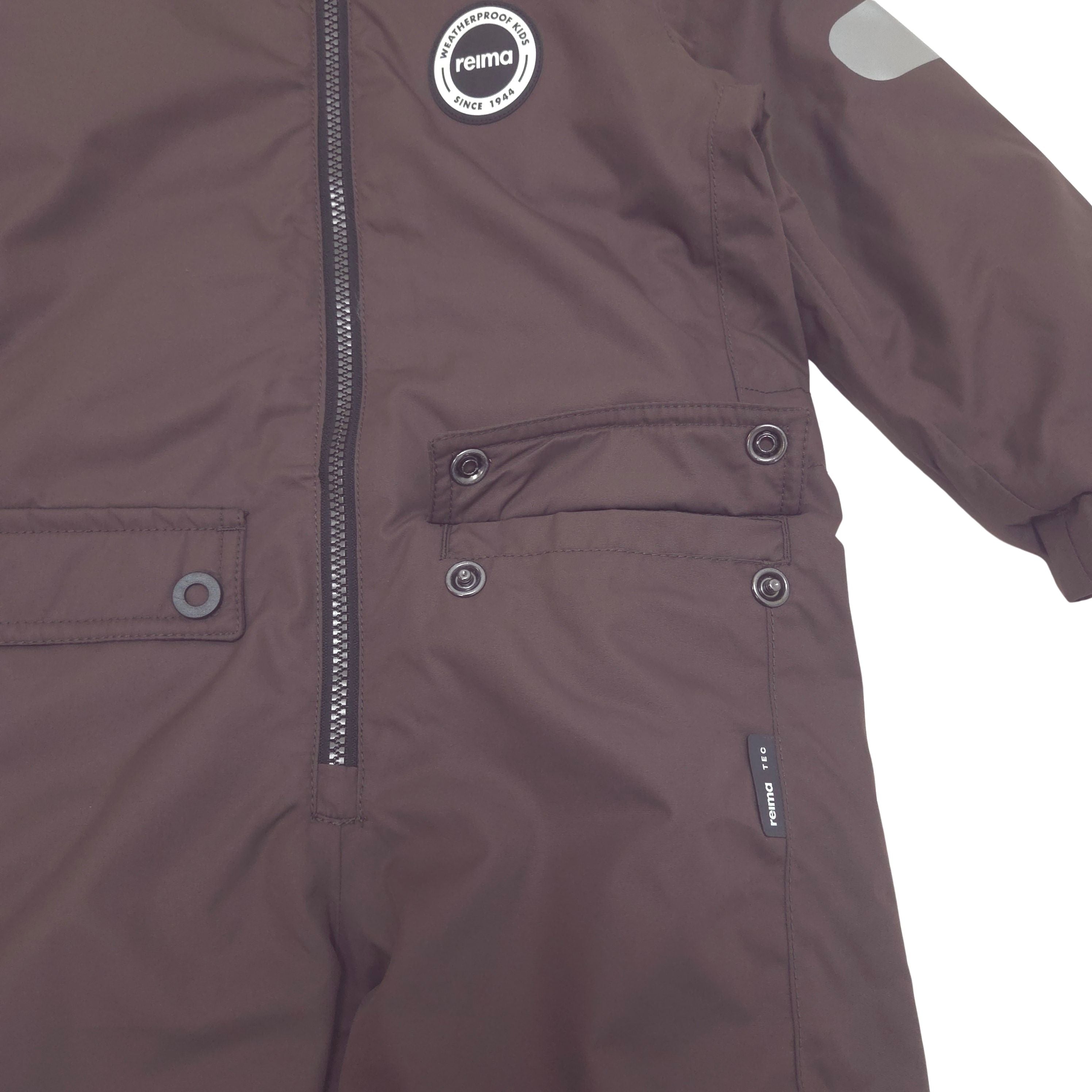 A close-up of a dark brown children's lined shell jacket by Rema, featuring a front zipper, buttoned flap pockets, and a circular brand patch on the chest.