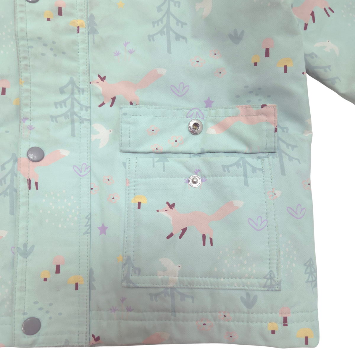 A close-up of a children's jacket or cardigan in a pale mint green fabric with a whimsical forest print featuring pink foxes, gray trees, and small mushrooms. The garment has a front snap-button closure and a large front patch pocket.