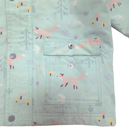 A close-up of a children's jacket or cardigan in a pale mint green fabric with a whimsical forest print featuring pink foxes, gray trees, and small mushrooms. The garment has a front snap-button closure and a large front patch pocket.